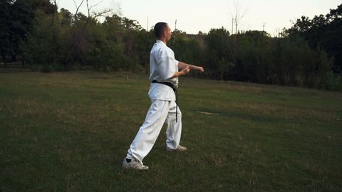 Adult Man in White Kimono and with Black Belt Trips Karate Beats in City Park at Dawn
