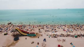 Aerial view of the beach filled with people on a hot sunny day. Sun umbrellas stand in yellow bright sand. People rest and sunbathe on the beach on a summer day near the ocean.