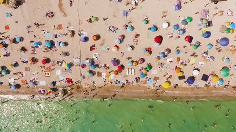 Aerial view of the beach filled with people on a hot sunny day. Sun umbrellas stand in yellow bright sand. People rest and sunbathe on the beach on a summer day near the ocean.
