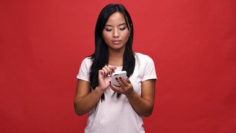 Surprised asian woman in t-shirt using smartphone and rejoice over red background
