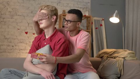 Pair of young multinational homosexuals sit on the couch, an African American presents a gift to his partner. Homeliness, gay, young LGBT family concept. 60 fps