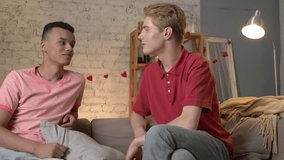 Couple of young multinational homosexuals sit on the couch, an American guy presents a gift to his partner. Homeliness, gay, young LGBT family concept. 60 fps