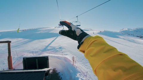 Man sits on ski lift going up mountain, shows shaka sign or hipster hang loose gesture on camera, plays with sun leak and beams, actuve lifestyle choice and leisure hobby winter activity