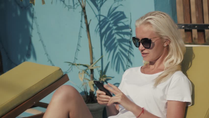 Joyful blonde girl checking mail in social networks while waiting friends near the hotel to swim in pool together. Adorable young woman in sunglasses sitting on chaise-longue and holding white phone