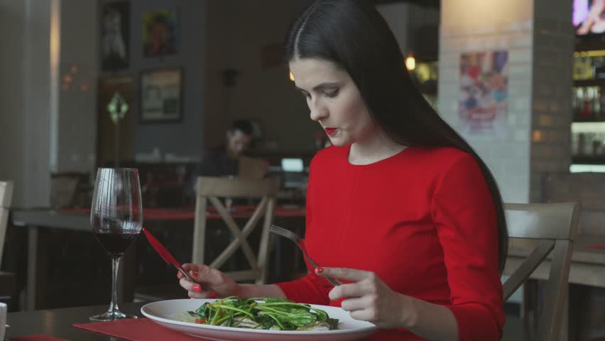 Young beautiful woman eating fish with vegetables and drinks wine in restaurant. The girl dressed in a red dress eats at restaurant.