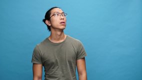 Confused asian man in t-shirt and eyeglasses with crossed arms looking around over blue background