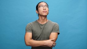 Pensive asian man in t-shirt and eyeglasses holding his chin and having idea over blue background