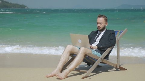 Young businessman is doing work using laptop while sitting on seashore. Bearded man is in work ing process, in deck chair on picturesque shore. Handsome guy in suit with tie has good time on beautiful beach
