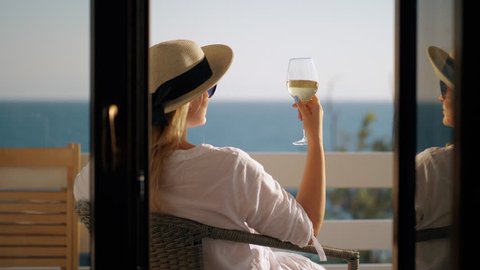 Cinemagraph - Young woman in hat and sunglasses having a rest with a glass of wine. She relaxing in arm-chair at the balcony overlooking the sea. Wind waving her hair