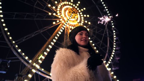SLOW MOTION. Happy excited woman in fur coat celebrating birthday and dancing in amusement park on the background of Ferris wheel and rides. Night and cold temperatures.