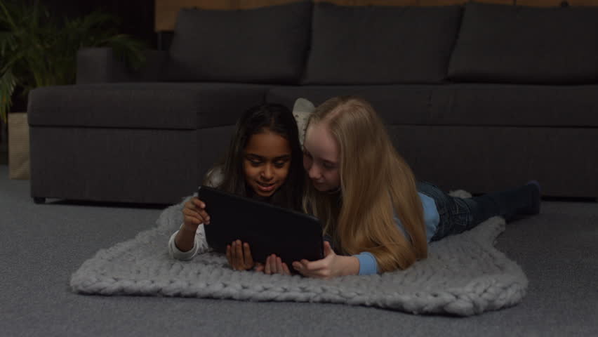 Adorable multi ethnic preteen girls browsing social networks on digital tablet while lying on the floor in domestic interior. 