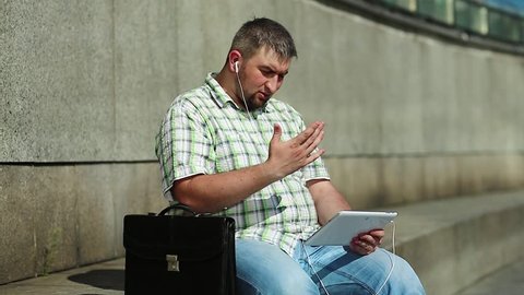 Businessman communicates via tablet computer. Man with tablet pc communicates through videochat. Fat man in checkered shirt with briefcase holds in hands tablet pc with earphones