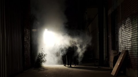 Wide shot of a silhouetted group of three men walking down a dark alley at night, passing through a backlit cloud of steam.