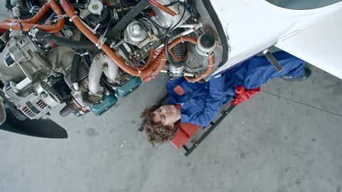 Zoom in from above of female aircraft mechanic lying on creeper underneath of airplane with exposed engine, repairing damaged part with screwdriver and then standing up and walking away