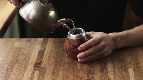 Close up of male hands preparing mate ethnic energetic drink Argentina Paraguay . man pouring water calabash gourd mate from metal kettle wooden table indoors process preparation shop advertisement.