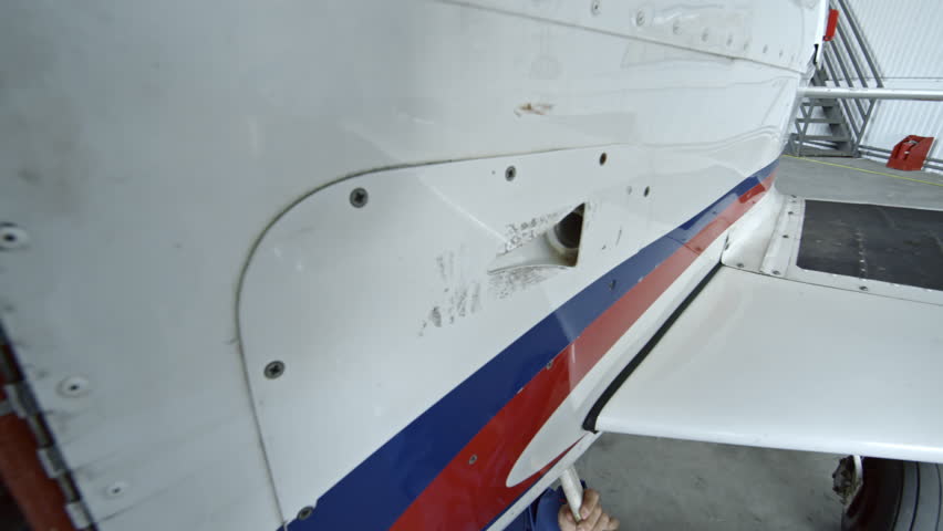 Young female aircraft mechanic in uniform lying on creeper underneath of jet airplane and wiping bottom with towel