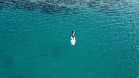 Aerial view of young girl stand up paddling on vacation. Tracking shot of a young woman SUP boarding