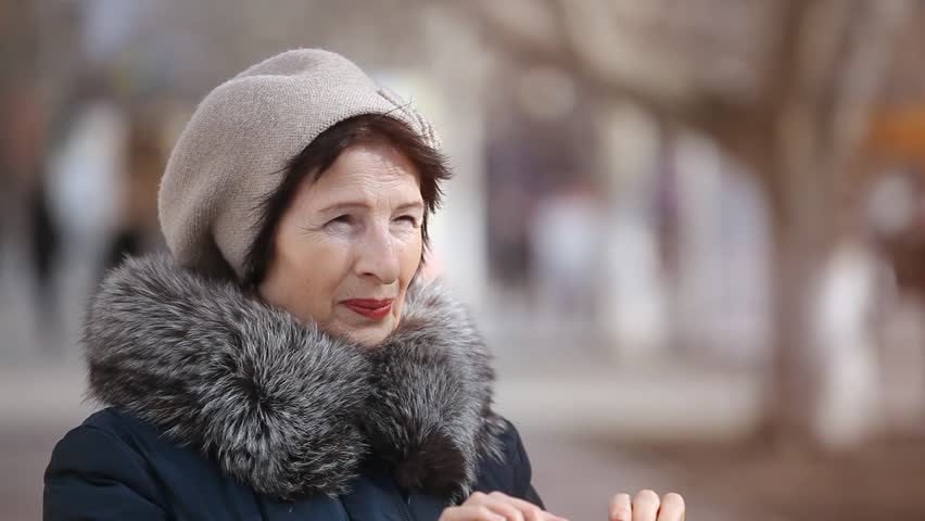   An impressive view of a brunette woman in a knitted hat and fur coat who snaps a photo in a city street. She stands in semi-profile in autumn 