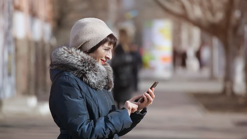 An impressive view of a middle-aged woman who stands in profile in a picturesque alley and touches the screen of her smartphone on a sunny day