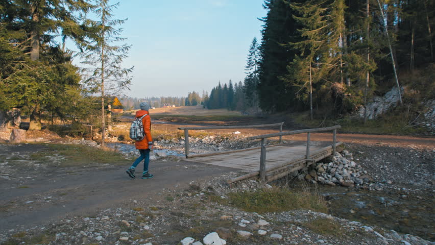 Teenager Girl with Backpack is Crossing a Bridge over a River in an Countryside in Autumn