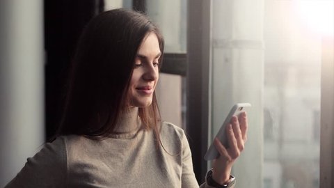 Slender brunette using the phone, wearing gray turtleneck top, indoor shot near the window: film stockowy
