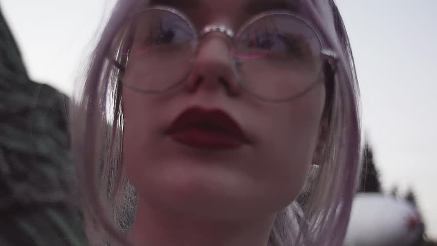Pretty blonde caucasian woman with pony tail and red lips, in round glasses at amusment park on summer evening, flashing lights, slow motion