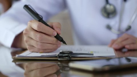 Doctor with stethoscope writing medical note with red heart decoration on table closeup blur background