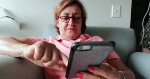 Older woman seated on sofa holding cellphone device. Candid 4K clip of elder woman touching smartphone screen