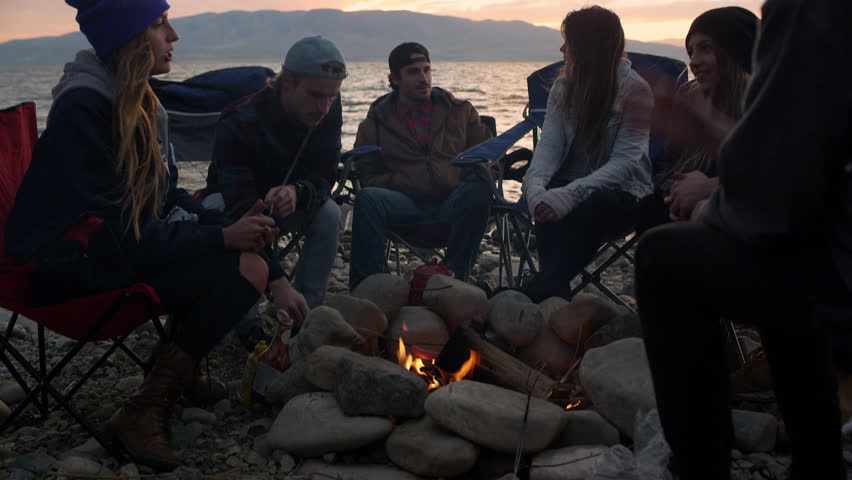 Group of family and friends gathered around campfire next to lake during sunset panning up to the sky.