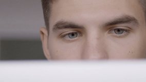 Portrait of young businessman who is sitting in the office and working on computer. The handsome man has grey eyes and is reading the report on the screen of his brand new laptop and his pupils are