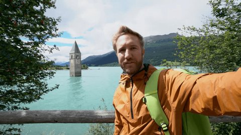 Travel male taking selfie at Lago di Resia, northern Italy.
Man takes a selfie in Resia lake, stunning mountain scenery, clock tower church in the background. Man taking selfies pov