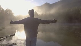 Young man standing by the lake at sunrise outstretches arms for freedom and positive emotions. Fog on the water surface, shot in 4K, Switzerland. People travel happiness positivity concept