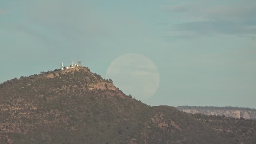 A pre-sunset super moon rising in a hazy cloud-streaked sky over a high desert landscape with a mountaintop fire tower and communications equipment; Tonto National Forest in Arizona