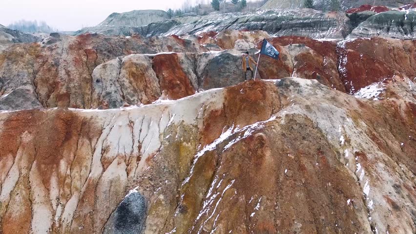 Man on top of a mountain with a flag in his hands. Footage. Successful silhouette man winner waving flag on top of the mountain peak, aerial view. Rear view man walking on top of rocky mountain peak