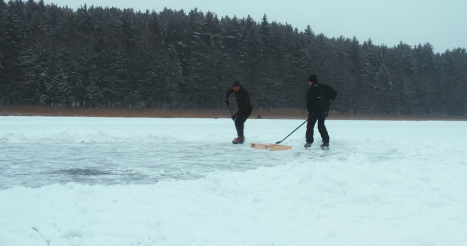 Two friends playing pond hockey on a frozen lake together, light snowfall. 4K UHD