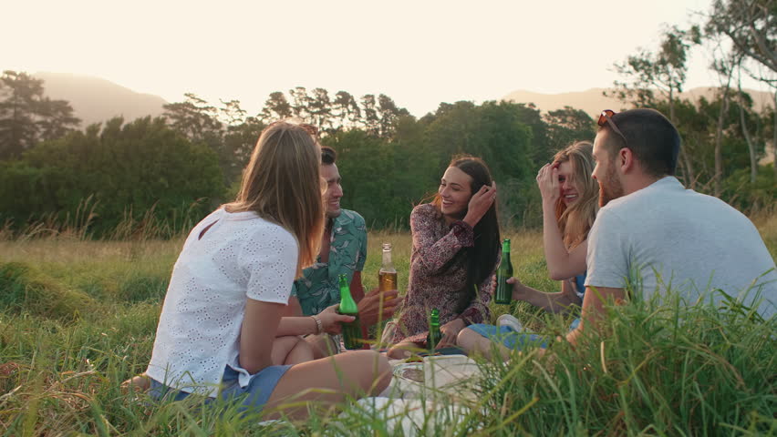 Friends relaxing together having a picnic at sunset, cheers drinks enjoying themselves