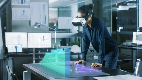 In the Office Professional Woman Wearing Augmented Reality Headset Interacts with Infographics Showing Statistics.  She Leans on the Table with Animated 3D Models Showing Company's Growth.