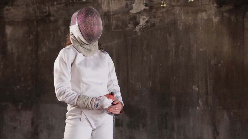 portrait of a young woman in a protective helmet, a lady stands against the background of an underground wall, a lady holding a rapier for sports competitions