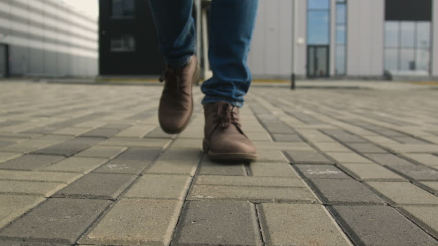 The one guy slowly confidence walks along the town footway in blue jeans and stylish brown leather shoe. Point of view from first person on legs close up. Autumn cold weather, pedestrian go up front Royalty-Free Stock Footage #1007405041