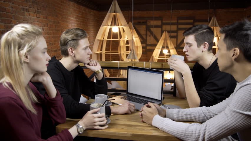 Group of people are communicating at table in cafe indoors. Men and woman talking while sitting at desk with coffee in cozy cafeteria. Three guys and lovely blonde speak actively and drink fragrant
