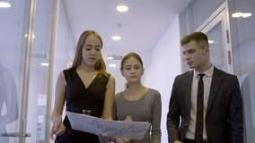 Three managers are walking through the office corridor and discussing digital currency market situation. Two businesswomen are holding papers with bitcoin graphs on and his male colleague in suit and
