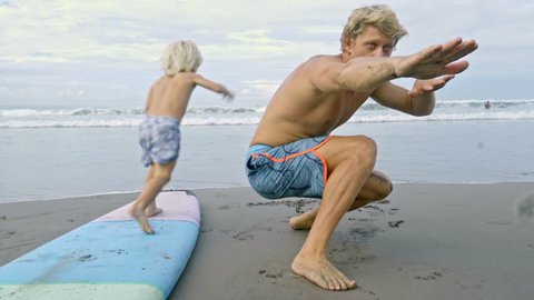 Father lying on stomach on beach teaching little son how to swim on surfboard and then kid running to ocean and playing with dog