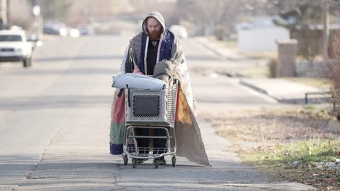 Homeless man pushing shopping cart down the street loaded with his belongings.
