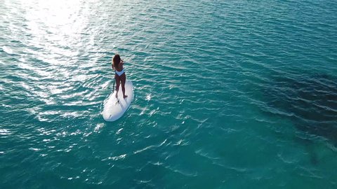 Aerial view of young girl stand up paddling on vacation. Tracking shot of a young woman SUP boarding