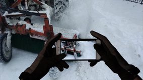 Monitoring and recording of the work of municipal services to clean the streets of snow. Winter, gloved hands hold a mobile phone and record to the video a moving tractor that removals the snow.