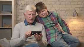 Grandfather and grandson are sitting on the couch using a smartphone, playing on a smartphone. Young fat boy and grandfather. Home comfort, family idyll, cosiness concept. 60 fps