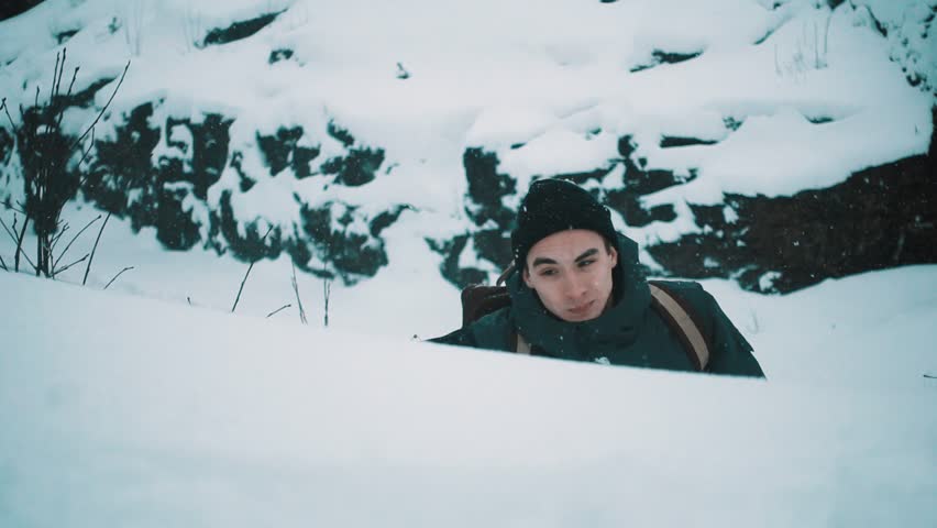 Young man in warm clothes and backpack holding canvas climbing mountain covered in snow on cold cloudy winter day, slow motion