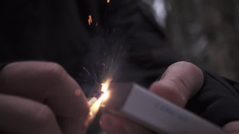 Male hand strikes match against box, ignites it on fire creates lots of sparks outside in woods, slow motion close up shot