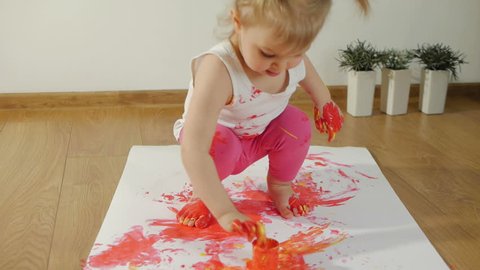 Little girl with smudgy paint fingers draws on a large sheet of white paper sitting on the floor