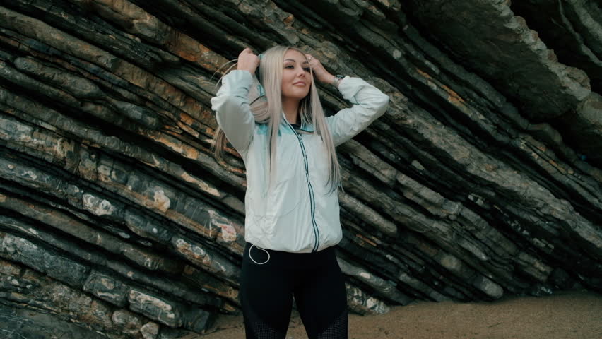 Young attractive woman with long blonde hair wearing a sports wear listens to music on the phone near winter sea. Slow Motion